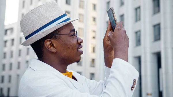 Smiling African American guy in white classic suit makes panoramic photo of highrise office buildings — Stock Photo, Image