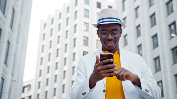 Афроамериканець в окулярах і капелюхах на смартфоні і посміхається з щастя від розмитих висотних будинків Стокове Зображення