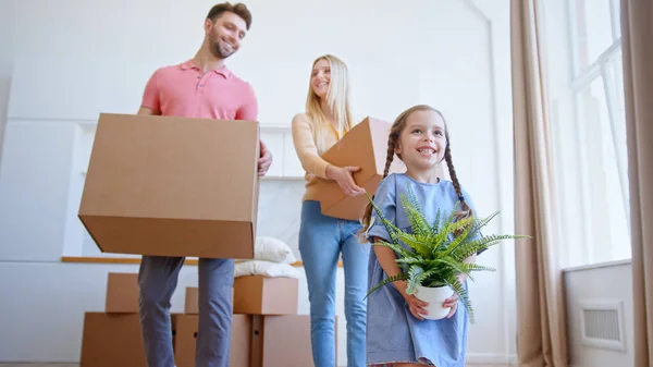 Funny little daughter helps parents with brown cardboard packages move holding small green pot plant in new flat — Stock Photo, Image