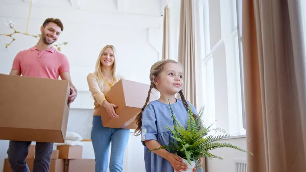 Rolig liten unge med långa flätor bär grön kruka växt mot föräldrar med stora kartongförpackningar — Stockfoto