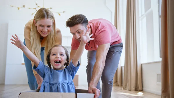 Joyful parents play with little funny daughter in blue dress moving kid along new apartment floor — Stock Photo, Image