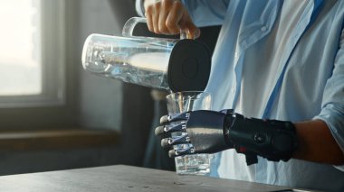 Yapay uzvu olan Cyborg adam mutfakta duran büyük kâse ve içeceklerden bardağa su döküyor.