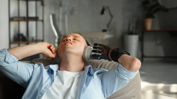 Relajado hombre discapacitado cyborg con la prótesis de mano bio negro escucha música en auriculares inalámbricos tumbados en un sofá grande —  Fotos de Stock