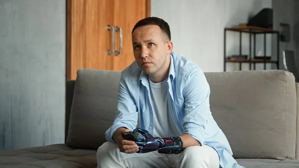 Interested bald guy with artificial hand prothesis switches television channels with remote controller — Stock Photo, Image