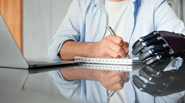 Company manager man with bio hand prothesis makes notes in paper notebook sitting at table — Stock Photo, Image