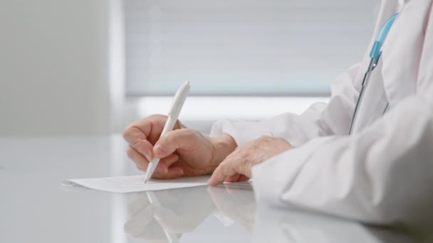 Medicul senior scrie pe bază de rețetă așezat la masă în clinică — Videoclip de stoc