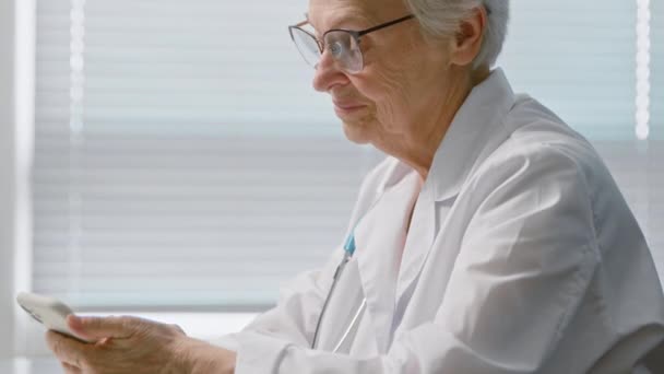 Dokter sms-bericht op mobiele telefoon zit aan tafel in kliniek — Stockvideo