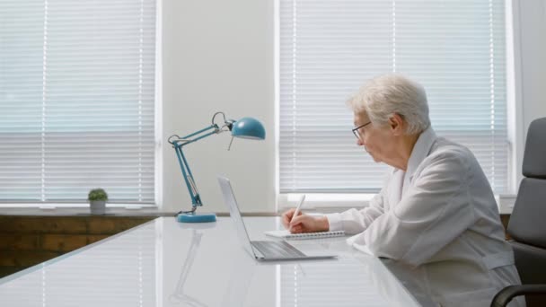 Зрелый доктор делает заметки, слушая пациента в видеочате — стоковое видео