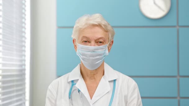 Vrolijke volwassen dame arts met grijs haar in jas met stethoscoop neemt wegwerp masker — Stockvideo