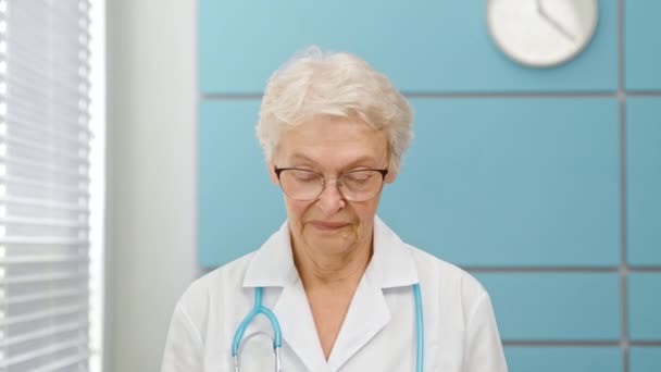 Senior short haired female doctor in white robe puts on disposable mask looking into camera — Stock Video