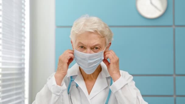 Χαρούμενη ώριμη γυναίκα γιατρός με γκρίζα μαλλιά στο παλτό με στηθοσκόπιο απογειώνει τη μάσκα μιας χρήσης — Αρχείο Βίντεο