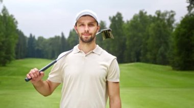 Golf sopalı genç adam golf sahasında kameraya bakıyor.
