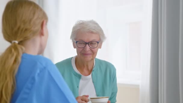 Starsza pani pije herbatę i rozmawia z młodą pielęgniarką w pokoju — Wideo stockowe