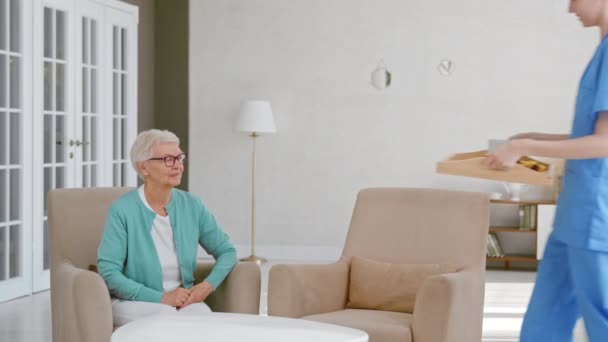 Verpleegster draagt dienblad met ontbijt naar senior vrouw in kamer — Stockvideo