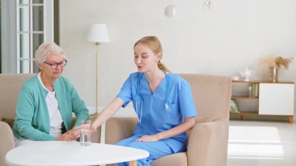 Joven médico da agua a la mujer mayor en la mesa en la habitación — Vídeos de Stock