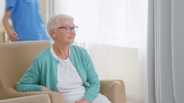Enfermera positiva viene a abrazar a la mujer mayor sentada en sillón — Vídeos de Stock