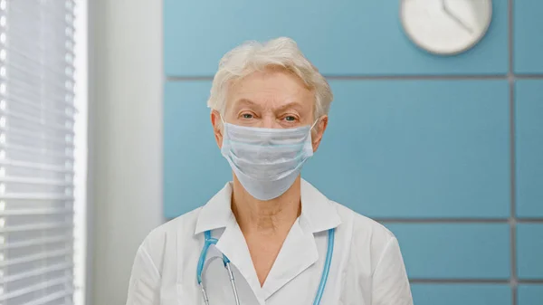 Medico anziano dai capelli grigi con maschera chirurgica e accappatoio bianco con stetoscopio guarda nella fotocamera — Foto Stock