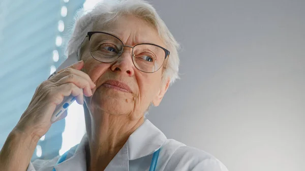 Médecin sénior joyeux avec lunettes parle sur téléphone portable lors de la consultation à distance par fenêtre — Photo