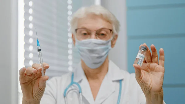 Gammal kvinnlig läkare med glasögon och engångsmask visar injektionsflaskan med vaccin och spruta mot kamera — Stockfoto