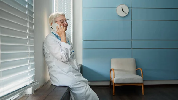 Happy mature lady doctor with glasses talks on cellphone with family member during at short break by window