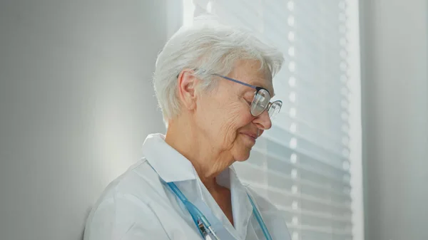 Médico fêmea madura feliz no manto branco com estetoscópio perto da janela no hospital claro — Fotografia de Stock