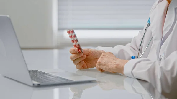 Woman doctor shows blister with small pink pills and explains prescription at videochat via laptop at table — Stock Photo, Image