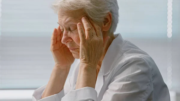 Уставшая старший врач в пальто массаж висков, страдающих от головной боли в удобном кресле — стоковое фото