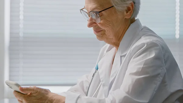 Alegre médico señora mayor con mensajes de texto gafas con receta en el teléfono celular sentado cerca de la ventana — Foto de Stock