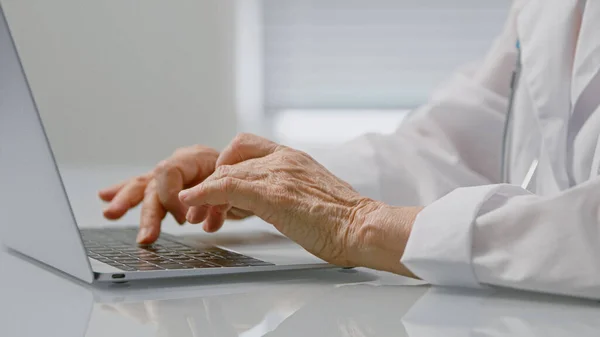Reife Ärztin tippt Text auf Laptop, sitzt am Tisch in geräumiger Klinik-Praxis und konzentriert sich auf faltige Hände — Stockfoto