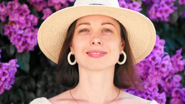 Chica joven feliz en un sombrero en flores lila mirando a la cámara — Vídeos de Stock
