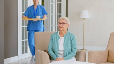 Mavi üniformalı sarışın hemşire kahvaltı taşır ve rahat koltuğunda hasta kadının yanına oturur.