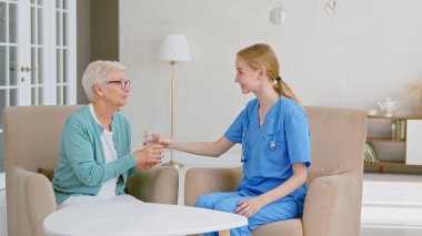 Üniformalı genç sarışın doktor, aydınlık odadaki küçük masada oturan yaşlı hasta kadına bir bardak su veriyor.