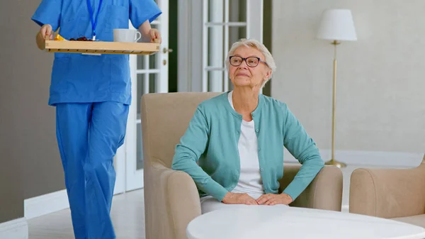 Caretaker in blue uniform carries tray with breakfast to mature grey haired lady sitting in armchair at table — Stock Photo, Image