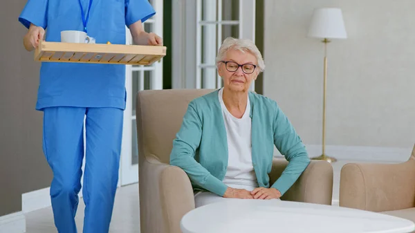 Lady operaio medico in uniforme blu porta bevande e frutta alla paziente anziana seduta in poltrona — Foto Stock