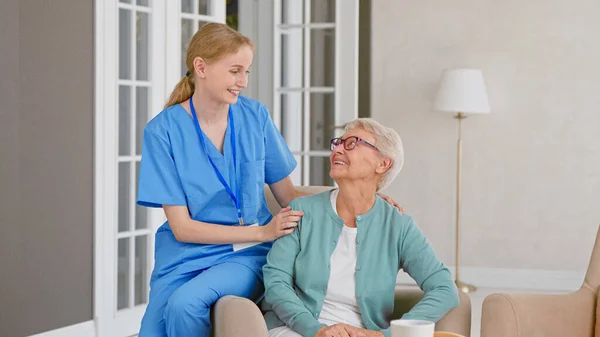 제복을 입은 어린 간호사는 편안 한 안락의자의 팔걸이에 앉아 있는 동안 노인 환자의 말을 듣는다 — 스톡 사진