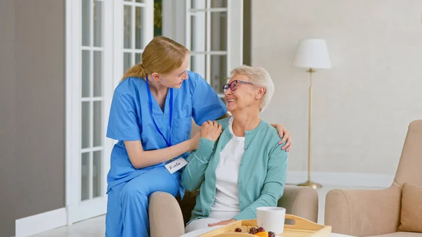 Uśmiechnięta blondynka pielęgniarka w mundurze dba o starszą kobietę siedzącą w wygodnym fotelu — Zdjęcie stockowe