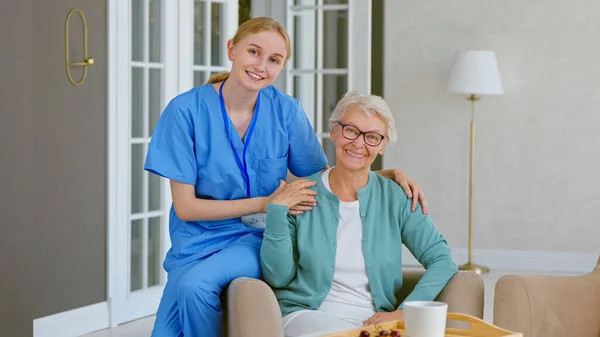 Усміхнена молода блондинка медсестра в уніформі піклується про старшу жінку, сидячи в зручному кріслі в світлій кімнаті Стокове Зображення