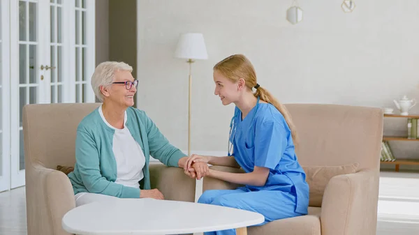 Positive doctor joins hands cheering up mature grey haired lady sitting in armchairs in stylish office — Stock Photo, Image