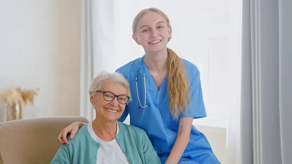 Mavi üniformalı pozitif hemşire rahat koltukta oturan yaşlı kadına sarılmaya geliyor. — Stok fotoğraf