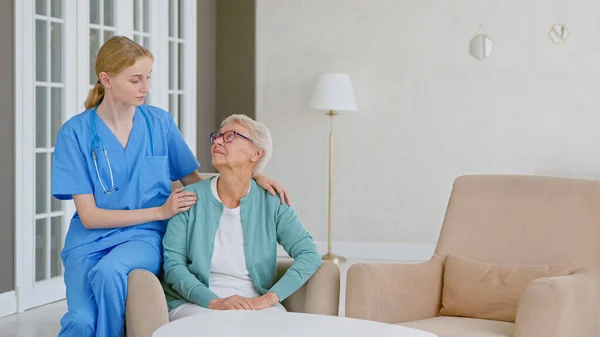 Enfermeira loira sorridente em uniforme cuida da mulher idosa na sala de luz na vida assistida — Fotografia de Stock