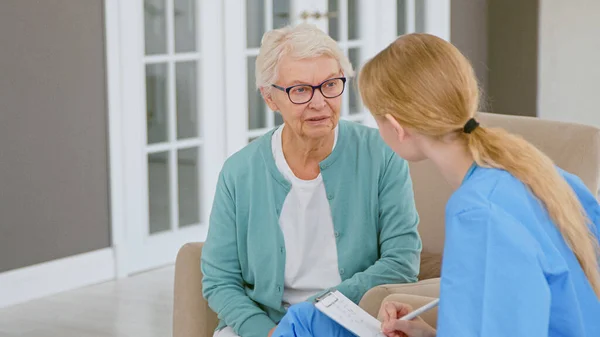 Junge blonde Ärztin interviewt kurzhaarige Seniorin, die sich bei Termin Notizen in Block macht Stockfoto