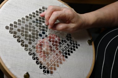 Beyaz keten üzerine ince bir geometrik dantel desenini dikkatlice dikerek nakış çemberine yerleştiren ellerin yakın plan görüntüsü. İnce iplikler ve kusursuz dikiş işi geleneksel bir iplik tekniğini gözler önüne seriyor. Sabır, işçilik ve...
