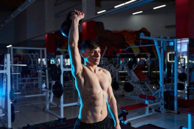 Çıplak gövdeli genç kaslı adam spor salonunda çalışıyor. Atletik erkek, çaydanlık zili ile egzersiz yapıyor. Spor, spor konsepti. Mavi arka ışık