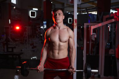 Spor salonunda kaslı bir adam halterle antreman yapıyor, kaslı erkek, çıplak karın kasları var. Kırmızı arka plan ışığı