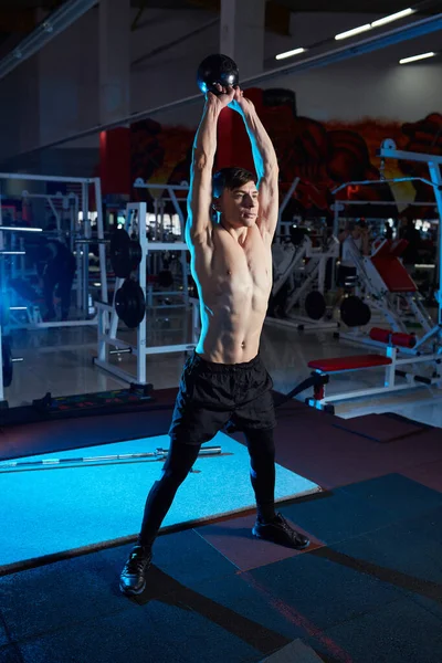裸の胴を持つ若い筋肉質の男がジムで働いています ケトルベルで運動する陸上競技男子大人 フィットネス スポーツの概念 青のバックライト — ストック写真