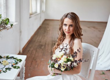 Parlak bir stüdyoda gelinlik içinde güzel bir gelin. Avrupa usulü düğün. Dekor ve buket elinde. Genç kız ayakta, oturuyor ve poz veriyor.