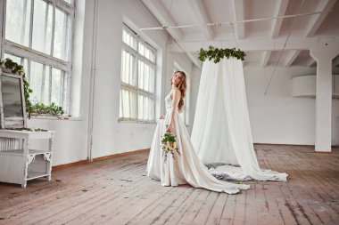 Parlak bir stüdyoda gelinlik içinde güzel bir gelin. Avrupa usulü düğün. Dekor ve buket elinde. Genç kız ayakta, oturuyor ve poz veriyor.