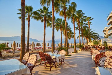 Şafakta plaj kafeteryasında. Kafede boş masalar, kumsalda güneşli bir sabah. Tahtadan masa ve sandalyeler