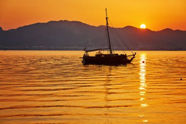 Deniz manzaralı dağ akşamı ve denizde küçük bir tekne. Gökyüzünde sarı güneş ışığı, günbatımı.