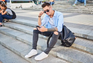 Genç bir adam, Avrupalı görünümlü, güneş gözlüklü ve siyah sırt çantalı. Kasaba meydanındaki basamaklarda elinde telefonuyla oturuyor ve çerçeveye bakıyor..